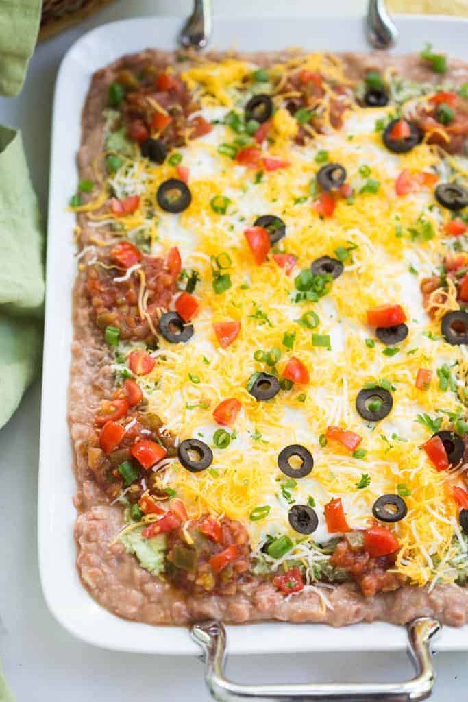 7-layer bean dip with refried beans, guacamole, sour cream, salsa, cheese, olives and green onion on a white serving tray.