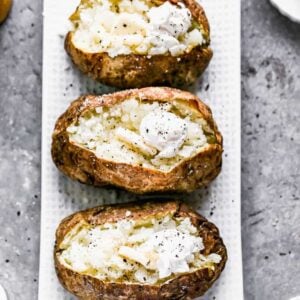 Three Air Fryer Baked Potatoes on a serving dish with the tops of the potatoes slit open and butter and sour cream added to the center.