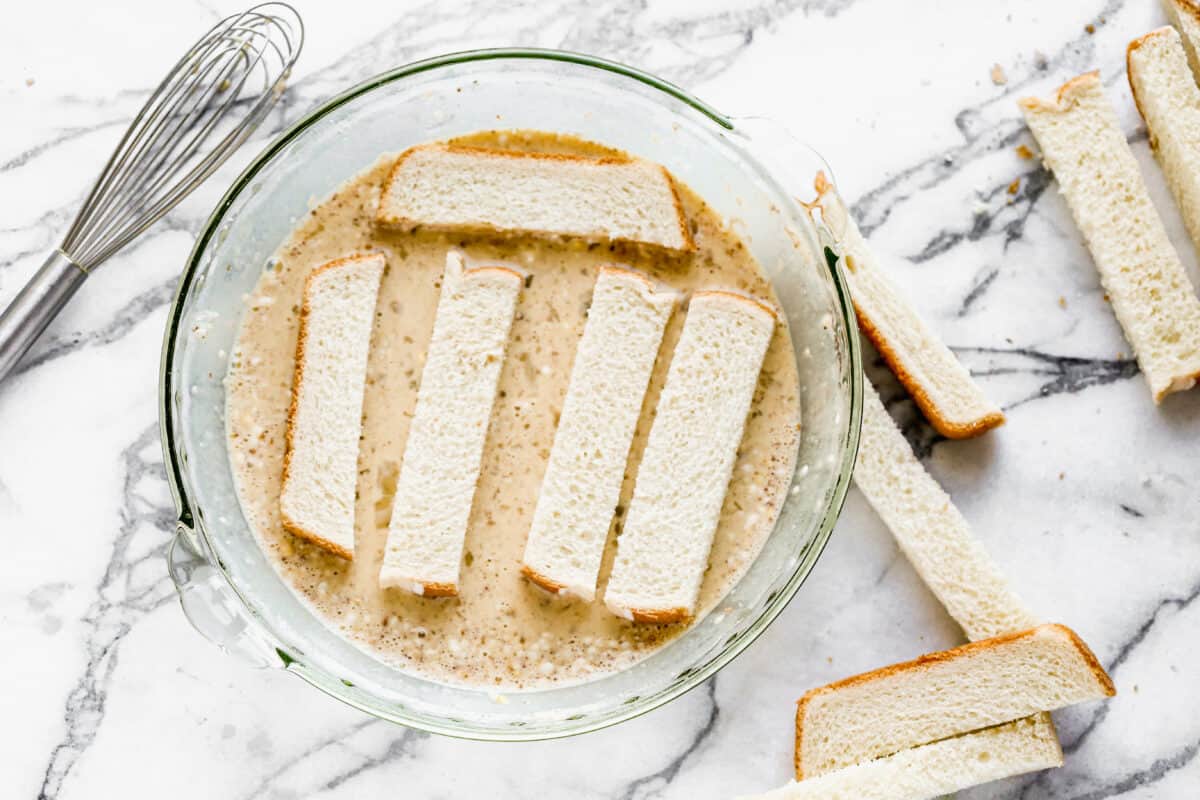 Strips of bread being dipped in a batter to show how to make the best french toast sticks.