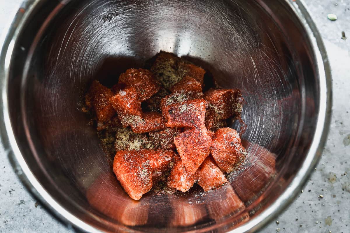 A mixing bowl with raw salmon bites and seasoning on top.