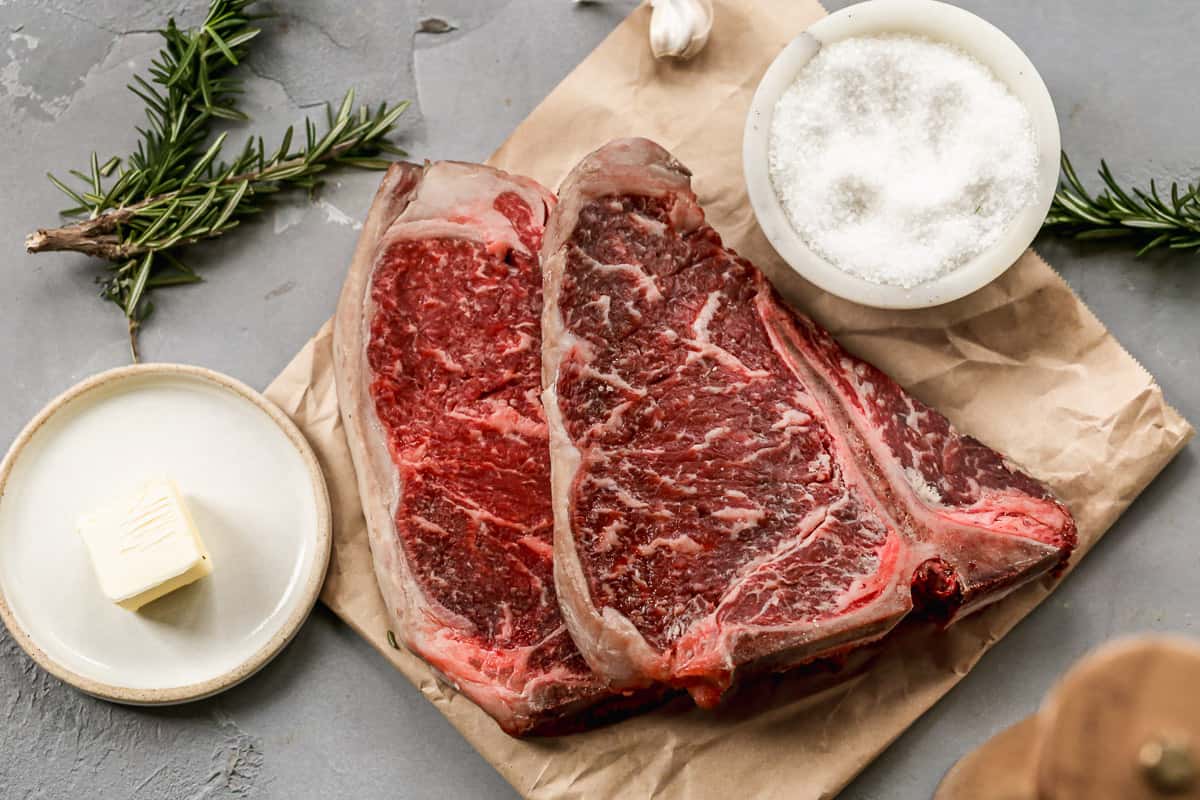 Two T-bone steaks on a piece of parchment next to butter, a clove of garlic, and a small bowl of salt, preparing to make air fryer steak medium rare.