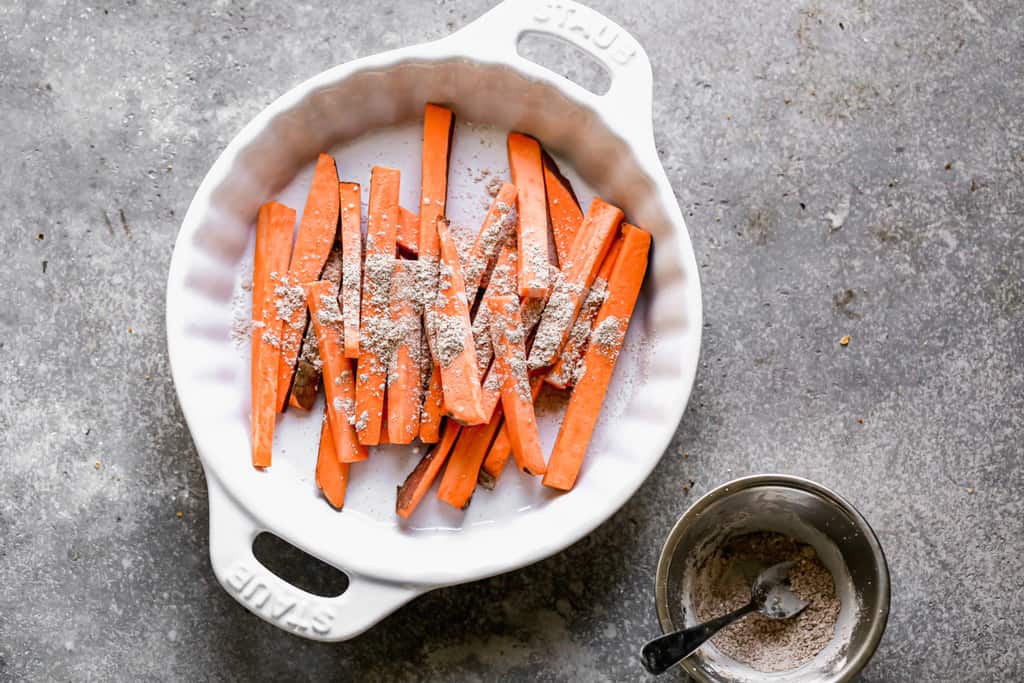 Thinly sliced sweet potatoes in a dish with seasonings sprinkled on top, to make fries.