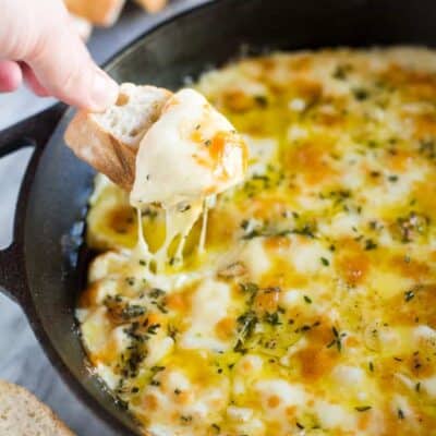 A cast iron skillet with baked fontina cheese dip, bread slices in the background and a hand taking a scoop out of the cheese dip with a bread slice.