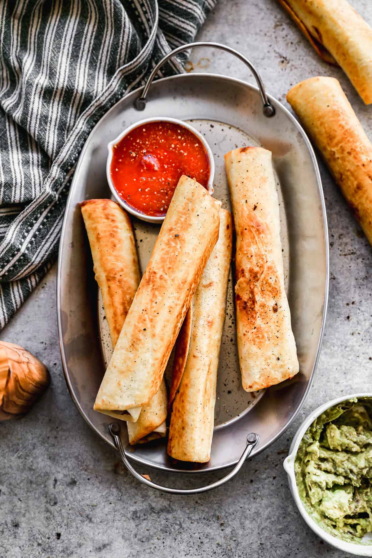 A platter of four crispy bean and cheese burritos with a side of salsa on it and a bowl of guacamole next to the platter.