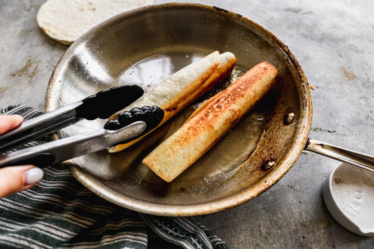 Two bean and cheese burritos being fried in a little oil in a pan to make it crispy.