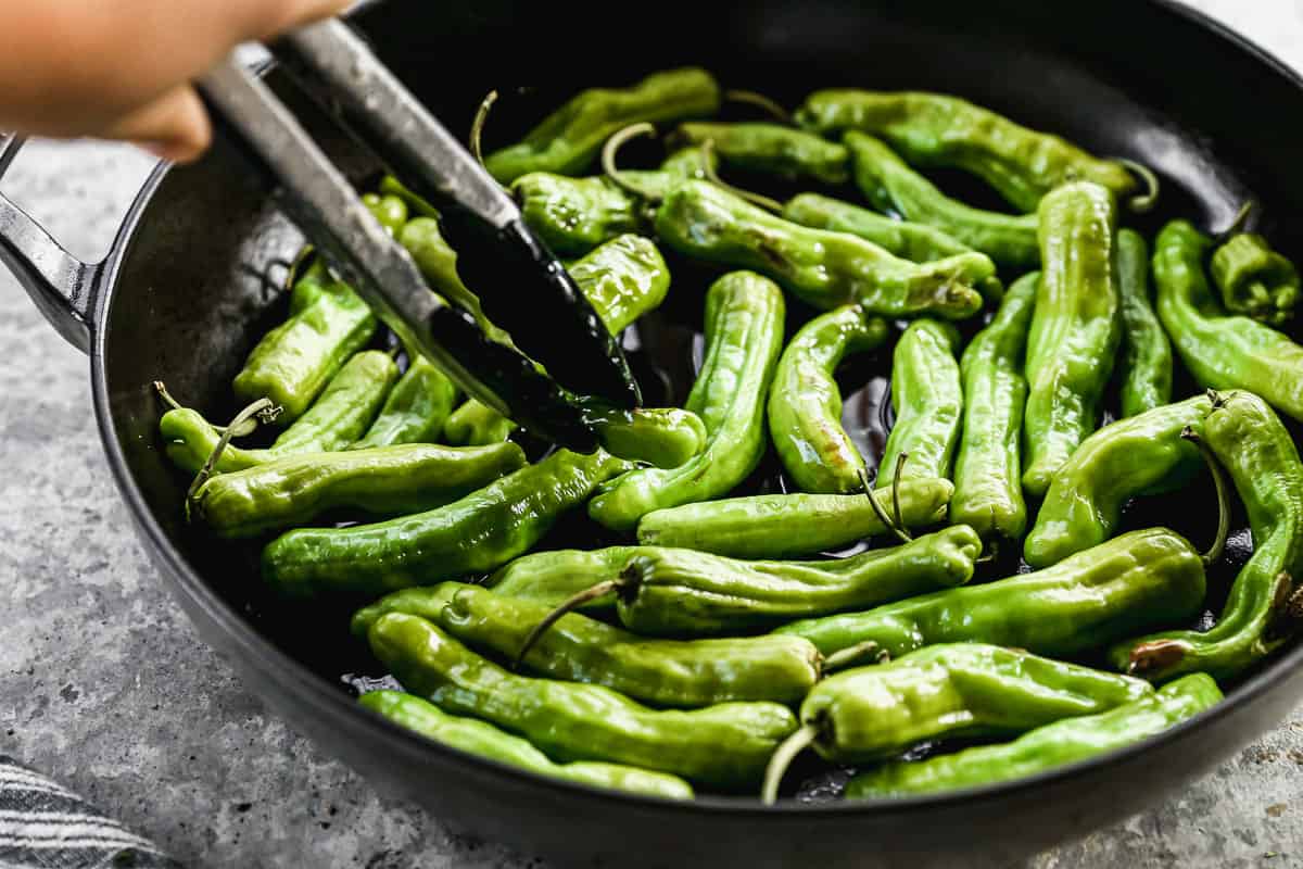 A close up image of tongs turning over shishito peppers to get them blistered on all sides.