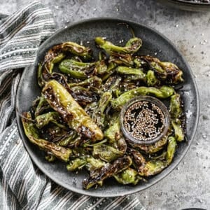 A plate filled with the best Blistered Shishito Peppers, drizzled with soy sauce, ginger, and honey, next to a sesame dipping sauce.