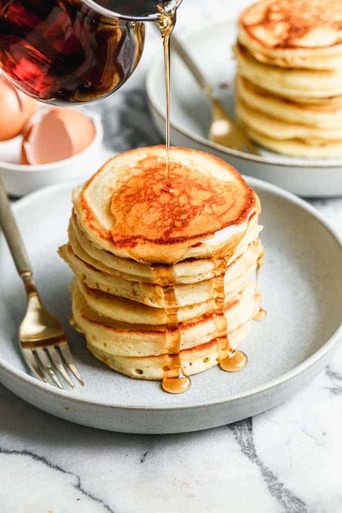 A large stack of buttermilk pancakes with syrup being poured over the top.