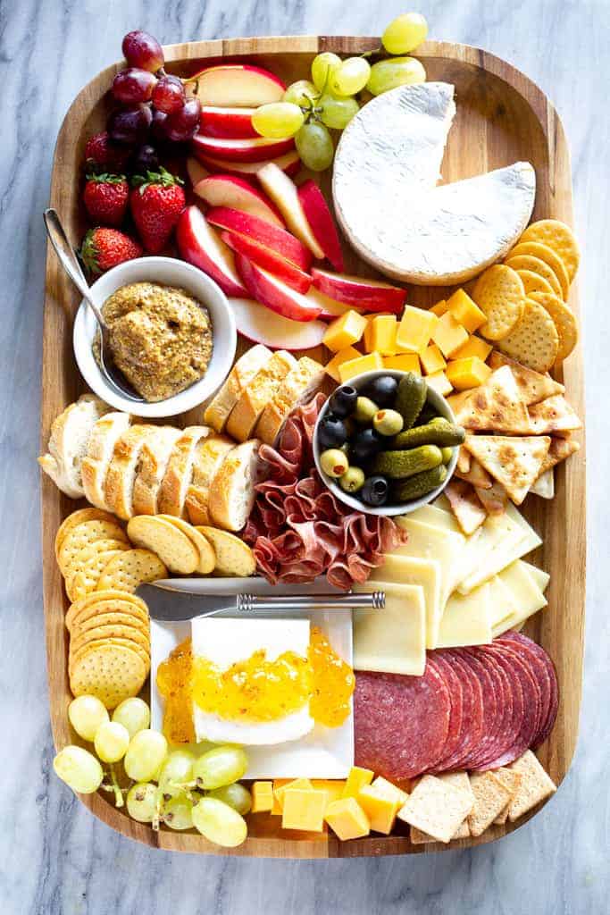 Charcuterie board with meats, sliced cheese, dips, crackers and fruit, served on a wood board.