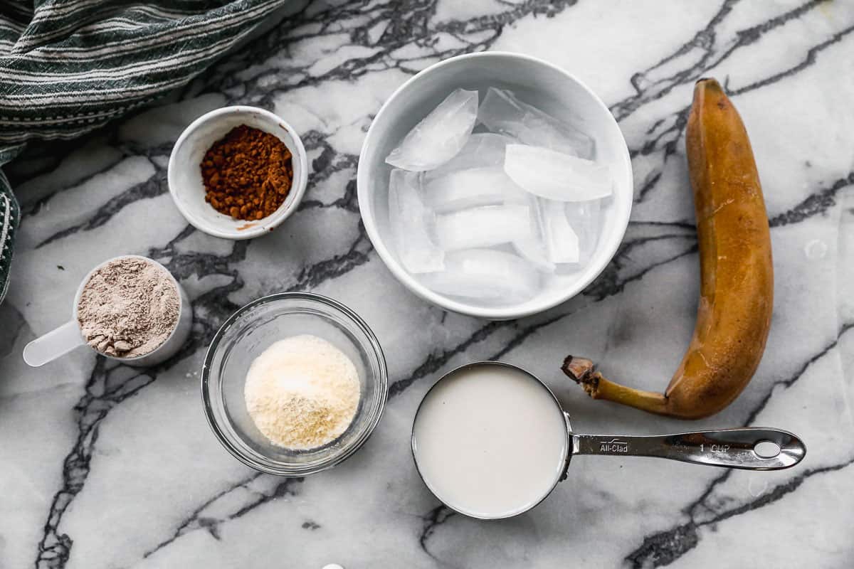 The ingredients needed for a Chocolate Protein Shake: chocolate protein powder, dark cocoa powder, almond milk, banana, ice cubes, and powdered peanut butter.