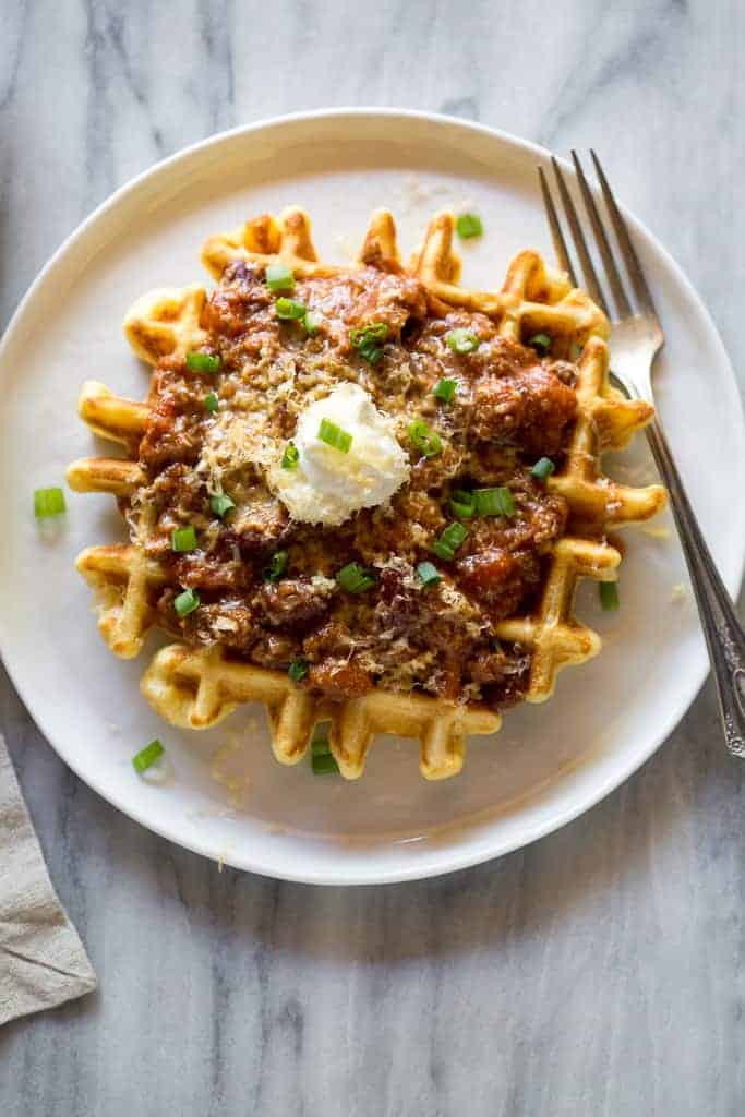 Cornbread waffle with chili and sour cream on it, on a white plate with a fork on it.