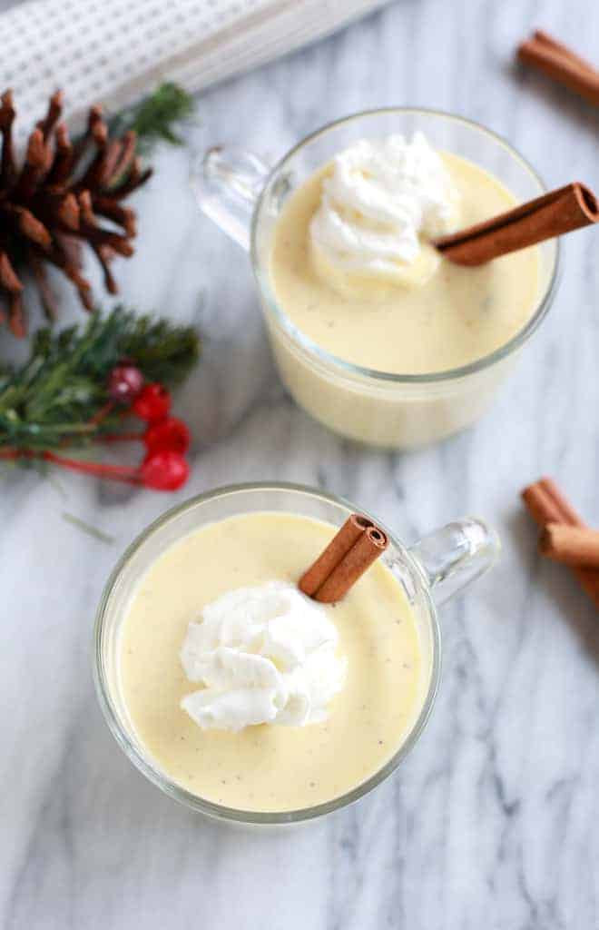 Overhead photo of two cups of eggnog with cinnamon sticks, pine, holly berries and pinecones.