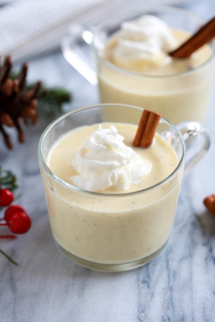 A cup of homemade eggnog served with whipped cream on top and a cinnamon stick, with holly berries, pinecones and another glass of eggnog in the background.