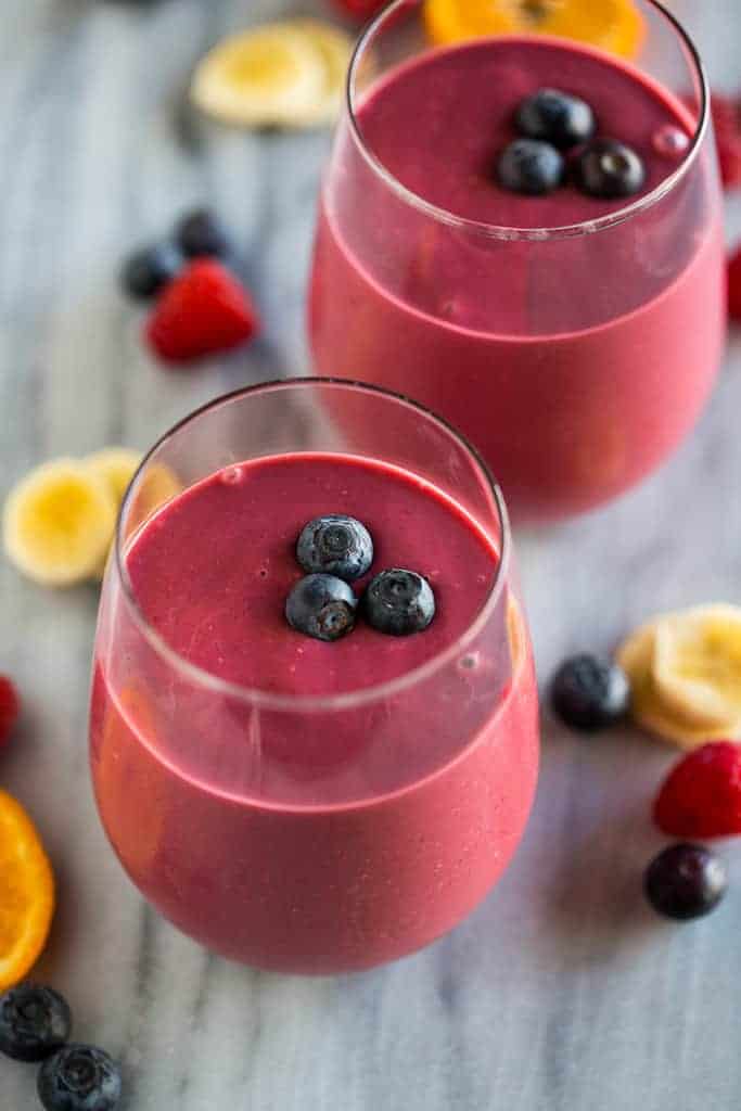 Two clear glass cups filled with a healthy breakfast smoothie with three blueberries placed on top and extra fruit in the background.