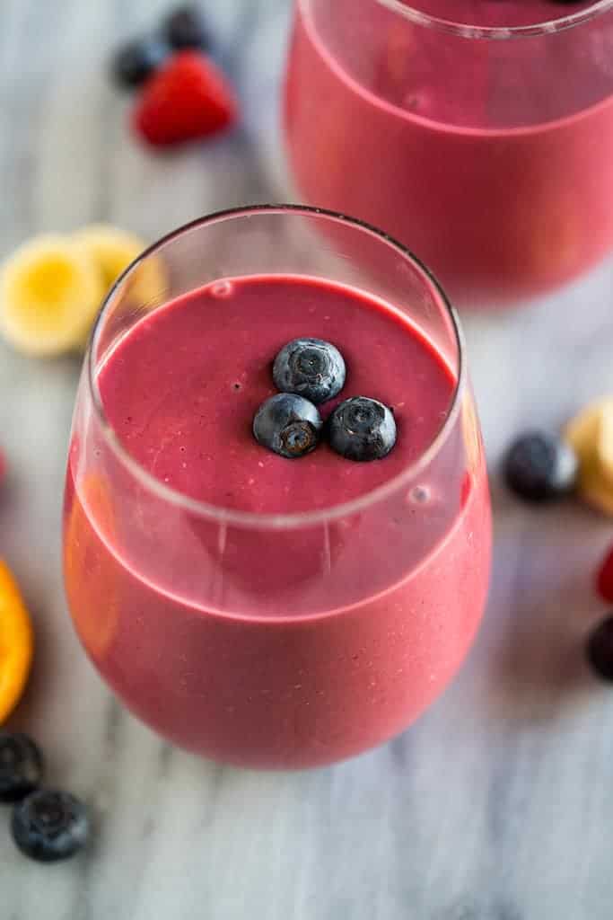 Close up photo of a healthy breakfast smoothie in a glass cup with three blueberries on top, orange slices, banana slices and blueberries in the background.