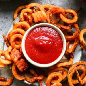 A small serving bowl of tomato ketchup on a plate surrounded by curly fries.