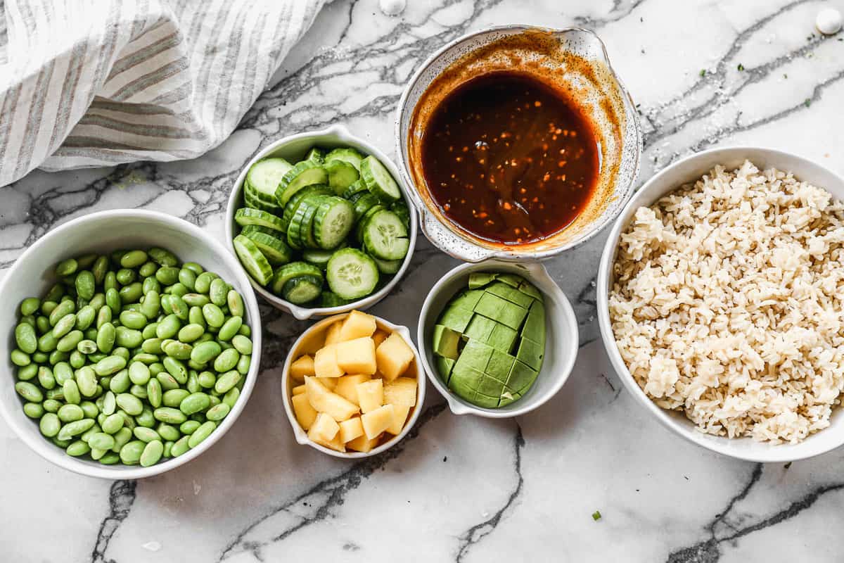 Jasmine rice, teriyaki sauce, chopped avocado, sliced cucumbers, mango, and edamame in bowls ready to assemble into easy salmon bowls.