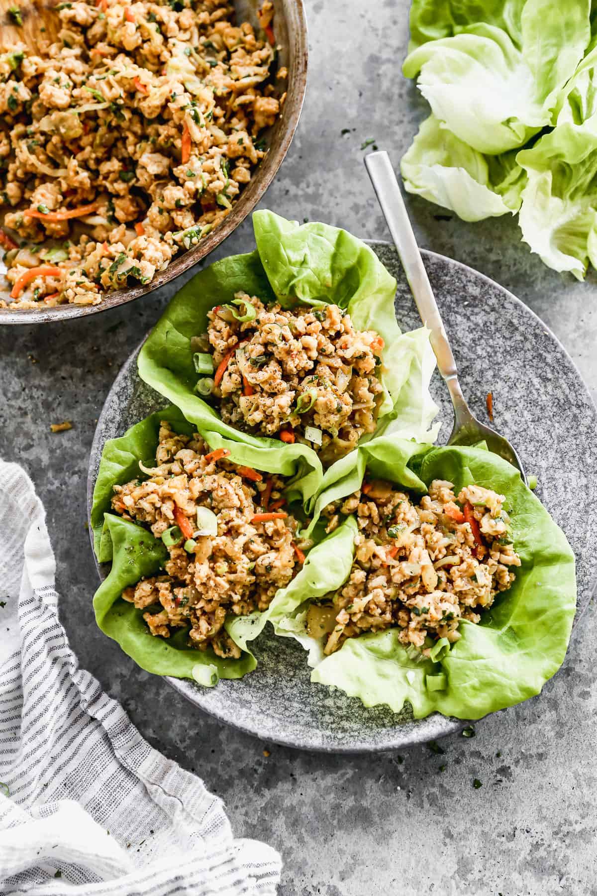 Three Thai Chicken Lettuce Wraps on a plate next to the pan of the chicken mixture and loose butter leaves on the counter.