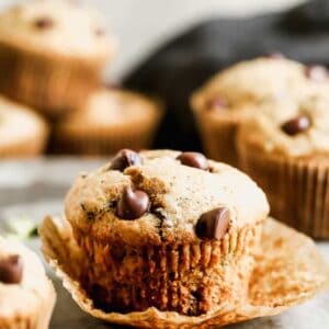 A zucchini muffin with its paper liner peeled down, surrounded by more muffins.