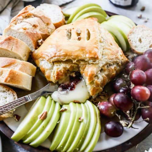 The best Baked Brie recipe wrapped in a puff pastry and served in the center of a platter surrounded by sliced baguette, grapes, and green apples.