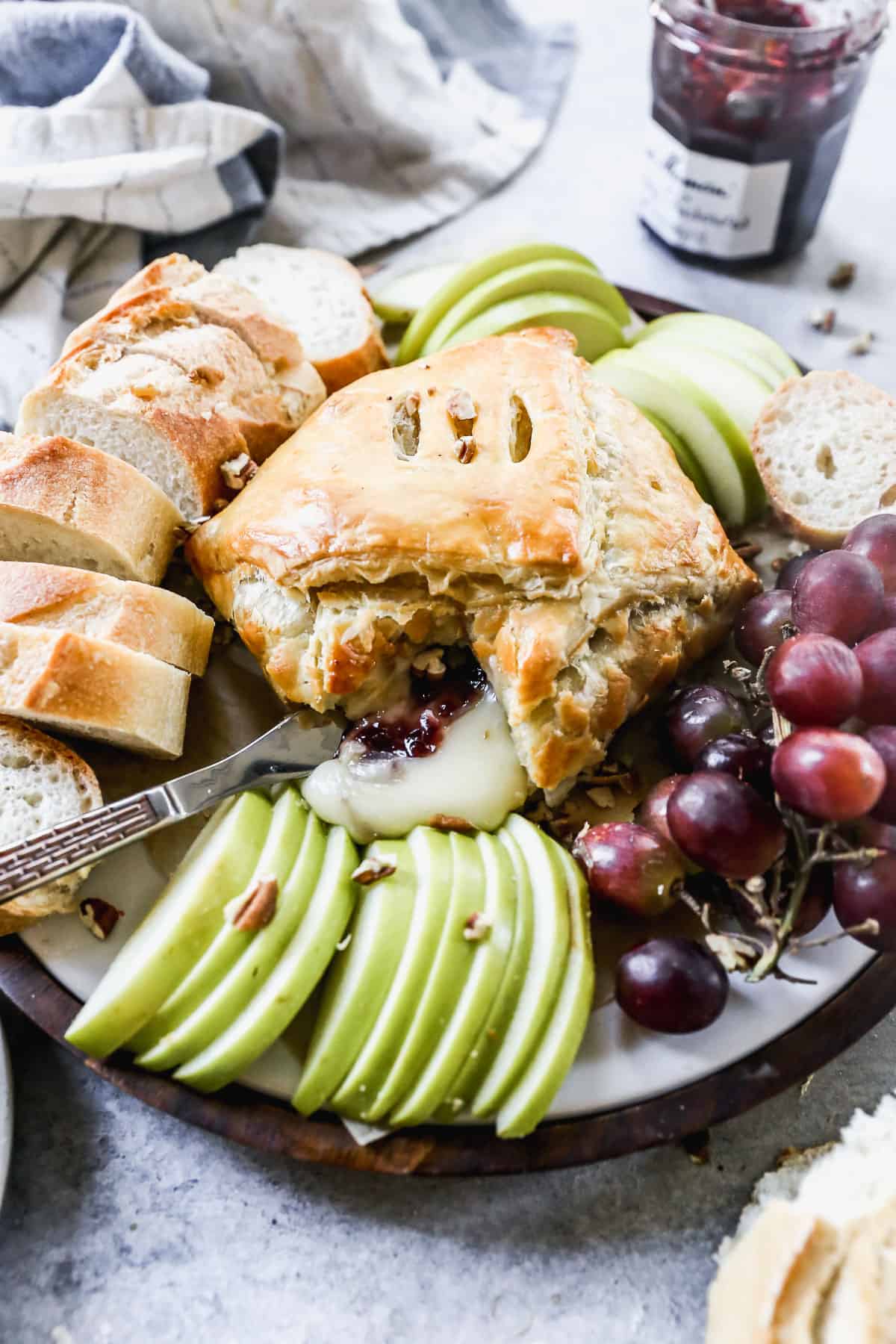 The best Baked Brie recipe wrapped in a puff pastry and served in the center of a platter surrounded by sliced baguette, grapes, and green apples. 