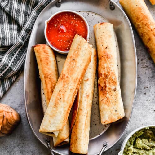 A platter of four bean and cheese burritos on a platter with a side of salsa and a bowl of guacamole.