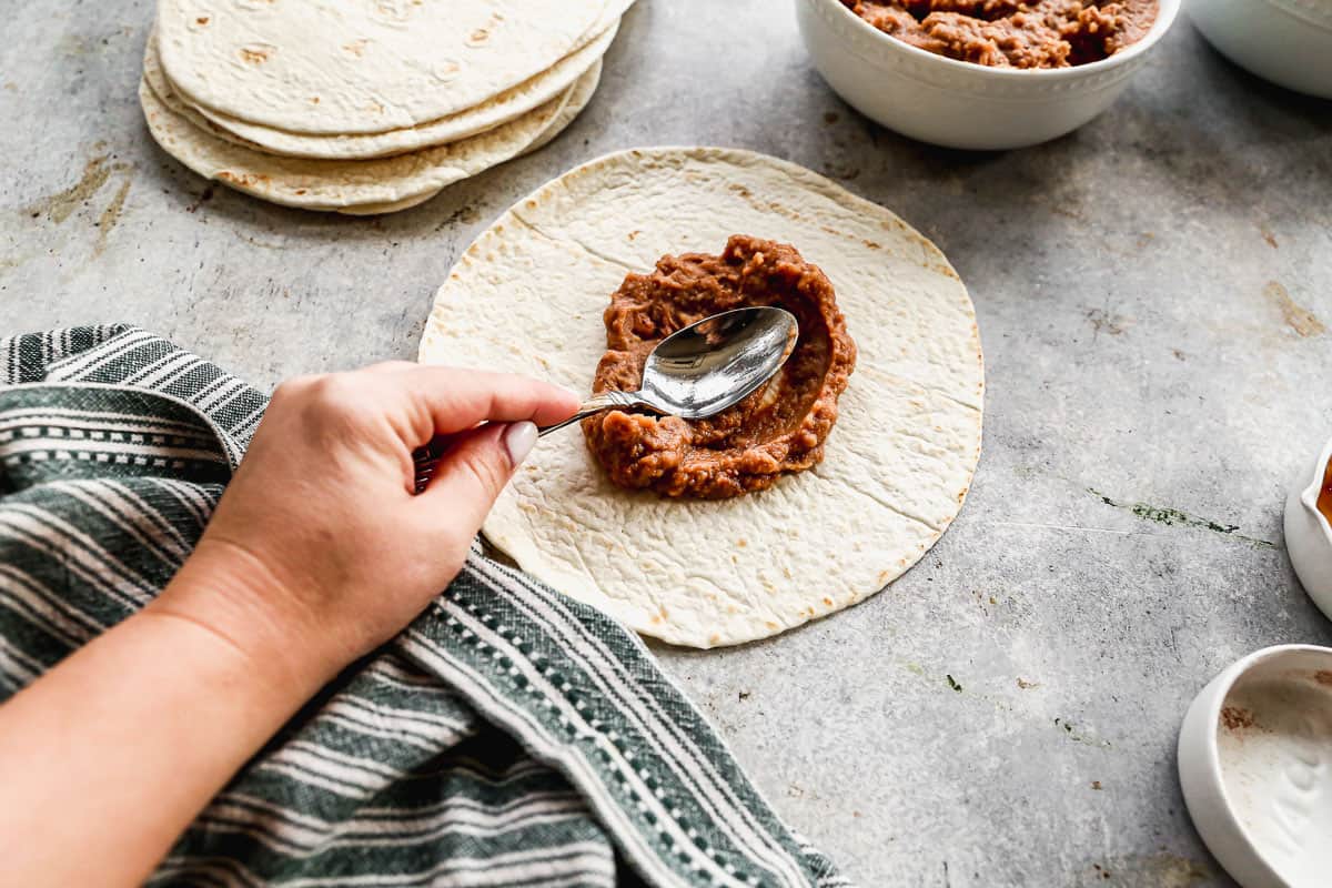 Someone spooning refried beans on a tortilla for homemade Bean and Cheese Burritos.