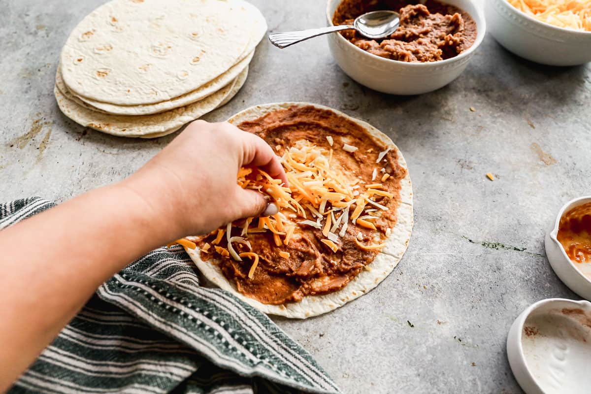 A Mexican cheese blend being spread on top of a tortilla with beans on it to make crispy bean and cheese burritos.