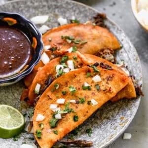 Birria Tacos served on a plate with a cup of consomé sauce for dipping.