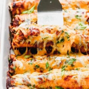 Beef enchiladas in a pan with a spatula lifting one out.