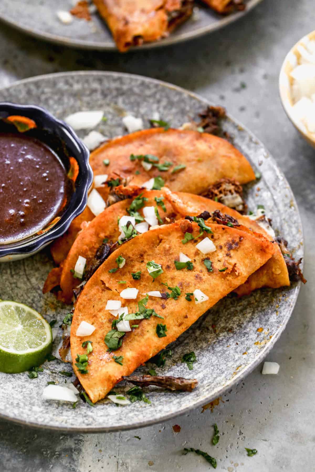 A plate with three of the best birria tacos served with a cup of consommé for dipping. 