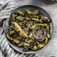 A plate filled with the best Blistered Shishito Peppers, drizzled with soy sauce, ginger, and honey, next to a sesame dipping sauce.