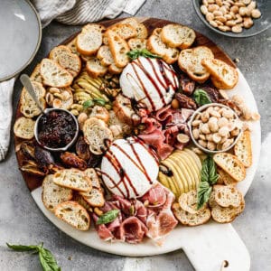 An easy Burrata appetizer drizzled with a balsamic reduction and arranged on a platter with mini toasts, jam, prosciutto, fresh pear, and dates.