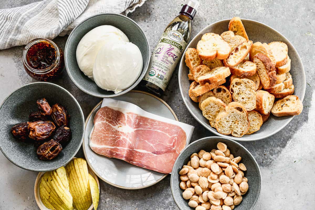 All of the ingredients needed to make the best burrata platter: burrata, balsamic reduction, mini toasts, nuts, prosciutto, fresh sliced pear, dates, and jam. 