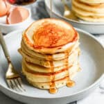 A stack of fluffy Buttermilk Pancakes on a plate with maple syrup being drizzled on top.