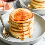 A stack of fluffy Buttermilk Pancakes on a plate with maple syrup being drizzled on top.