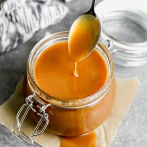 A homemade Caramel Sauce recipe in a glass jar with some caramel dripping off a spoon to show the smooth consistency.