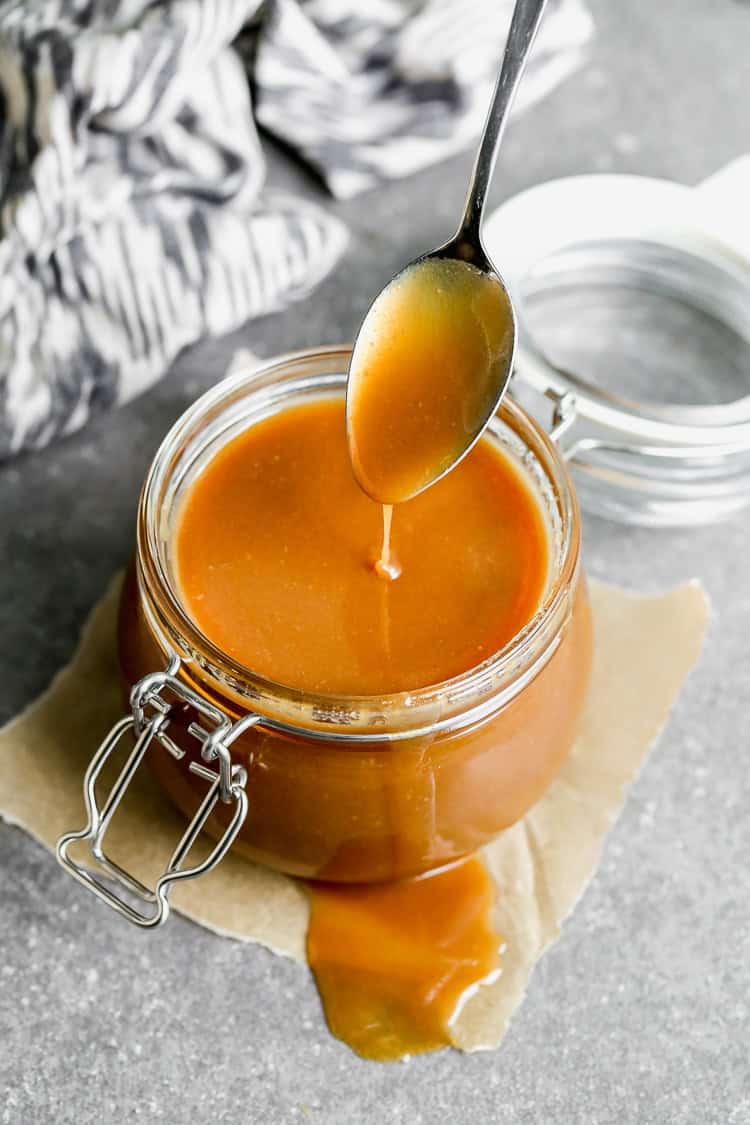 A homemade Caramel Sauce recipe in a glass jar with some caramel dripping off a spoon to show the smooth consistency. 