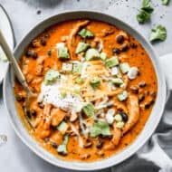 A bowl of a creamy Chicken Enchilada Soup recipe topped with chopped avocado, shredded cheese, and a scoop of sour cream.