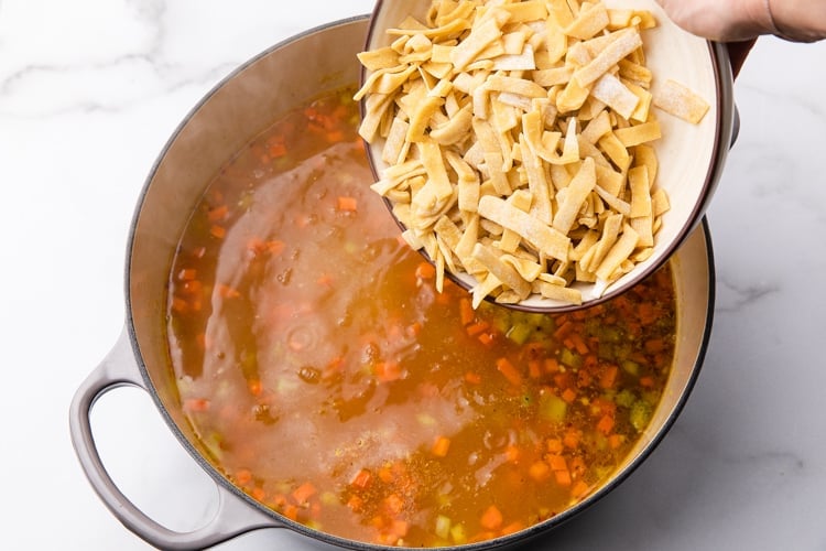 Homemade egg noodles being added to a pot of chicken stock and veggies.