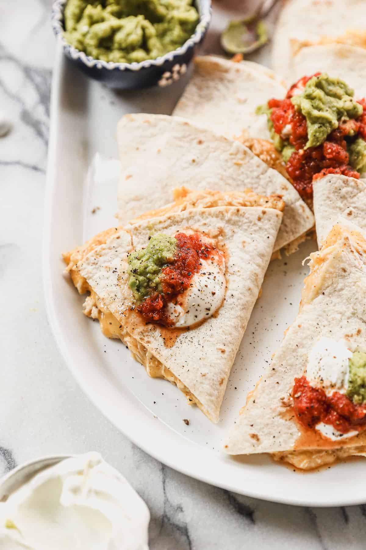 A crispy homemade Chicken Quesadilla sliced in wedges and served on a platter with guacamole, salsa, and sour cream.