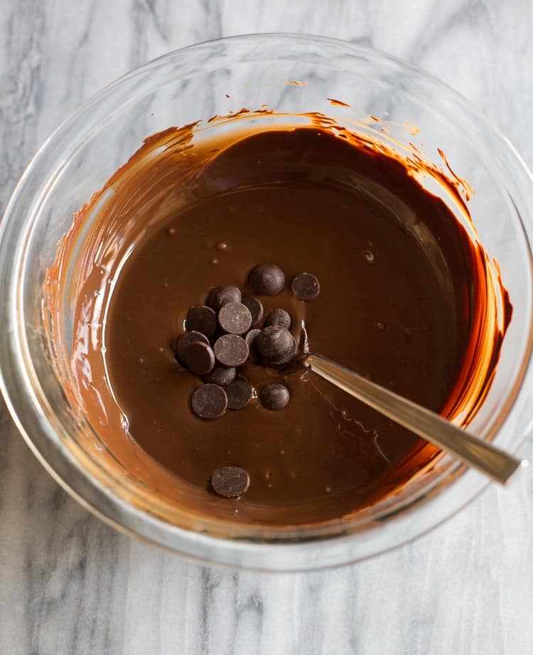 Overhead photo of a clear glass bowl with melted chocolate in it and a few bittersweet baking chips added to the bowl with a spoon to stir them in.