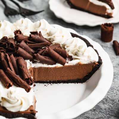 An easy Chocolate Cream Pie recipe with one piece on a plate, showing the oreo crust, chocolate filling, and whipped cream topping. Chocolate curls are placed on top.