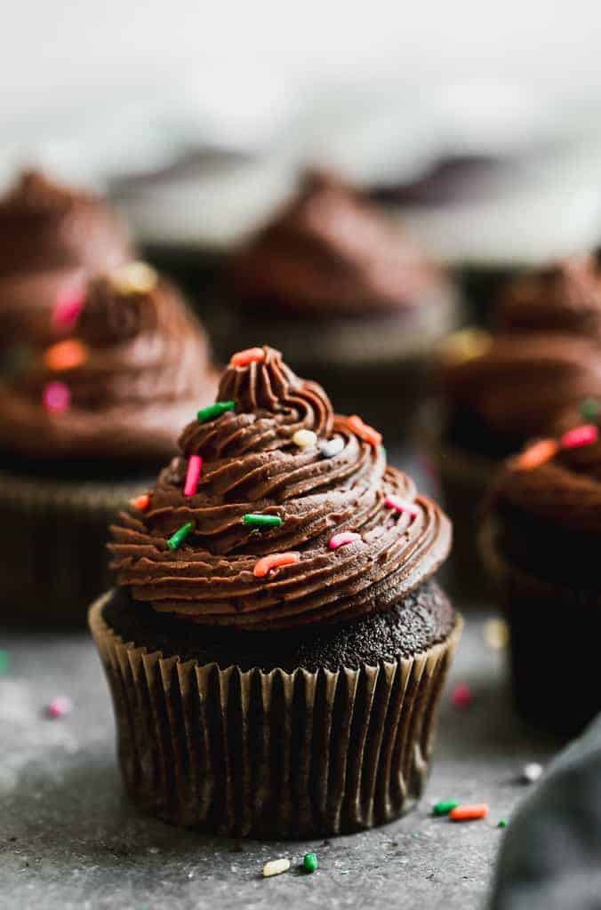 Chocolate cupcakes with buttercream frosting and sprinkles on top, lined up on a grey board . 