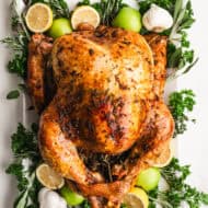 An oven baked Turkey covered in a garlic herb butter and roasted to perfection. It's placed on a hwite platter with fresh herbs, green apples, and lemons.