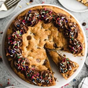 A chocolate chip cookie cake recipe with chocolate frosting swirled around the edge, topped with rainbow sprinkles.