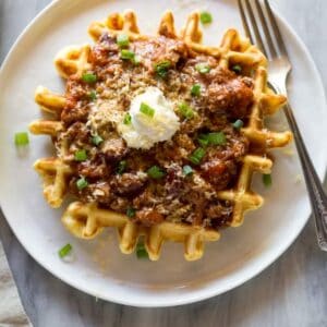 Cornbread waffle with chili and sour cream on it, on a white plate with a fork on it.