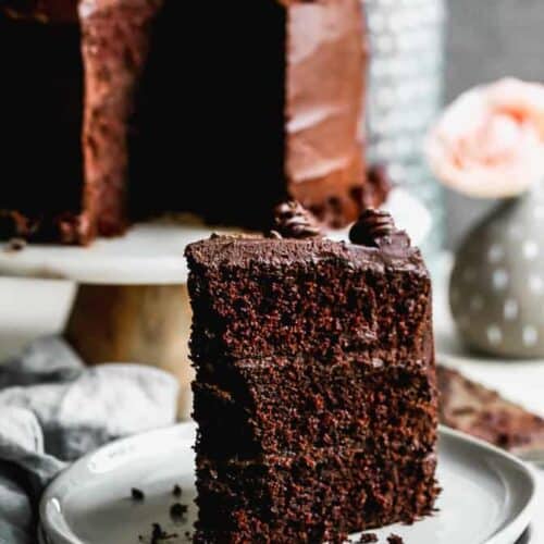 A slice of dark chocolate cake on a white plate and the remaining cake in the background.