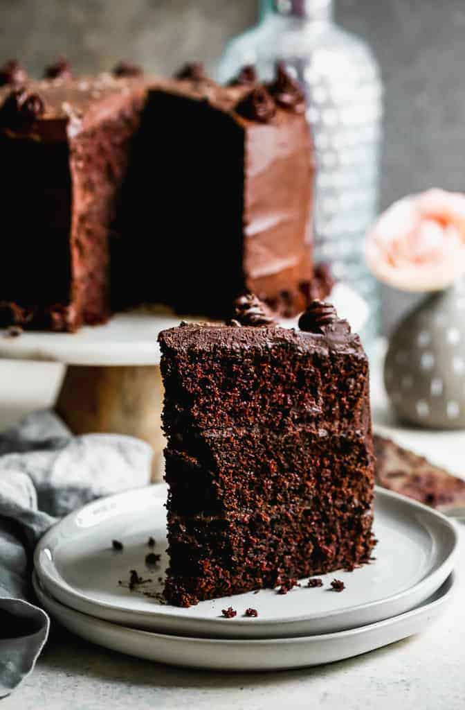 A slice of dark chocolate cake on a white plate and the remaining cake in the background.