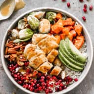 A harvest bowl recipe with tender chicken, brussels sprouts, sweet potato, pecans, pomegranate seeds, and a sliced avocado on top of a bed of coconut rice.